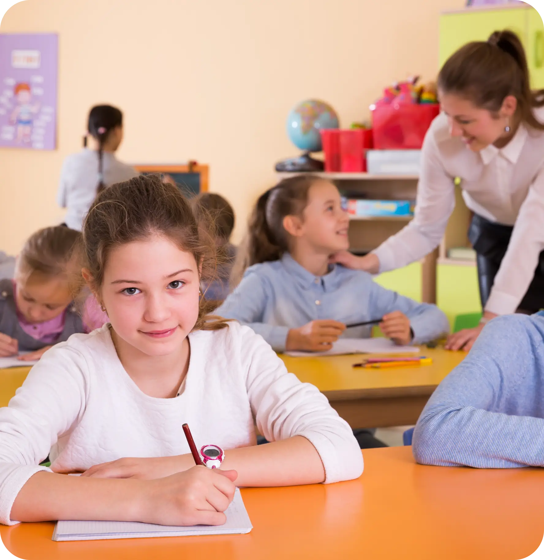 A girl sits at a desk writing in a notebook while other children work and a teacher interacts with students in a classroom.